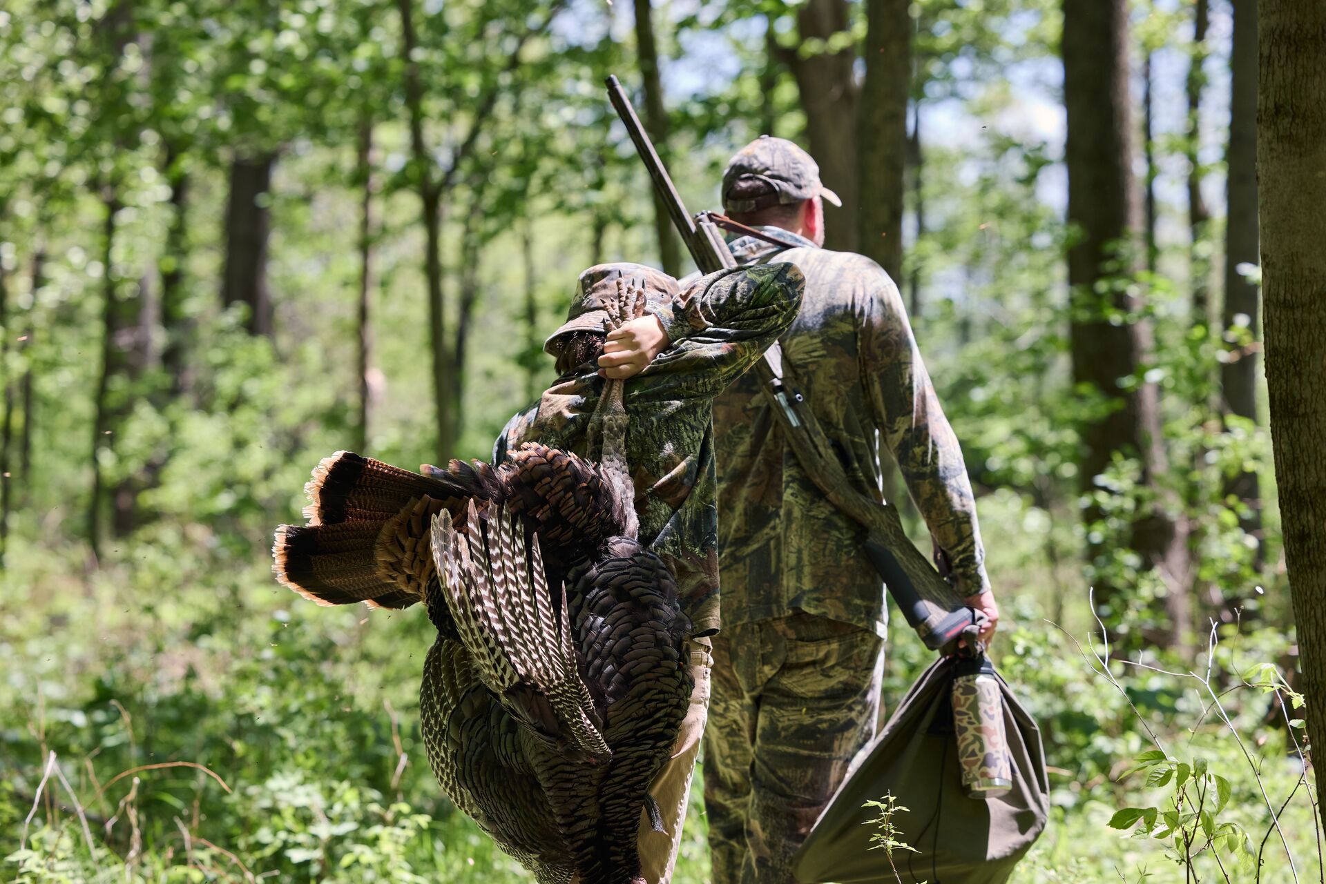 Boy carries turkey while walking behind adult after hunt. 