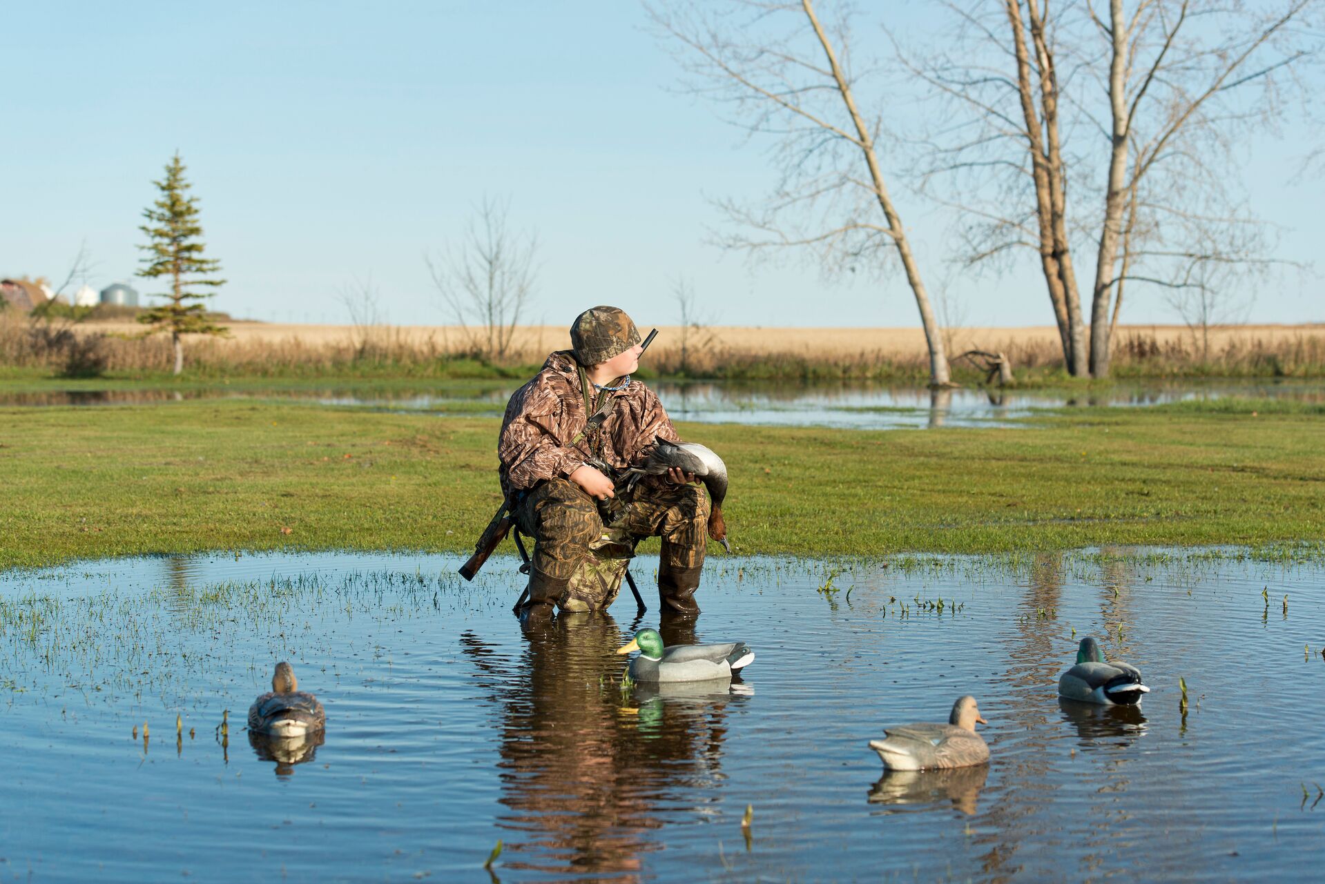 Boy in camo with firearm and duck decoys in pond. 