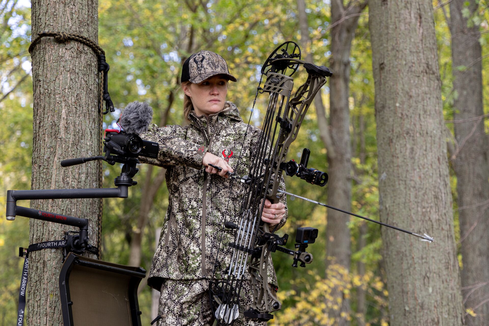 A woman wearing camo in a tree with a compound bow for hunting.