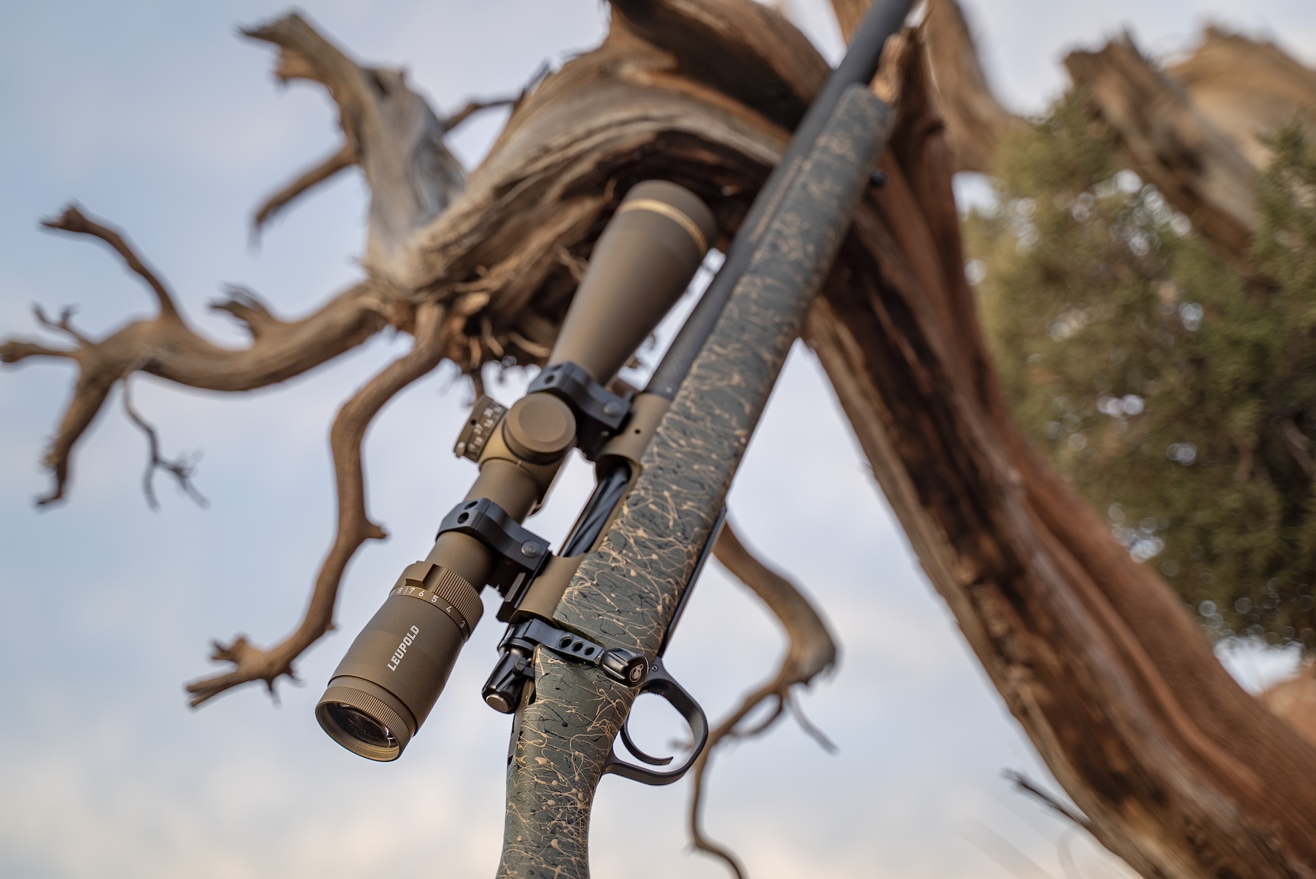 Bolt action rifle in camo leans against a tree. 