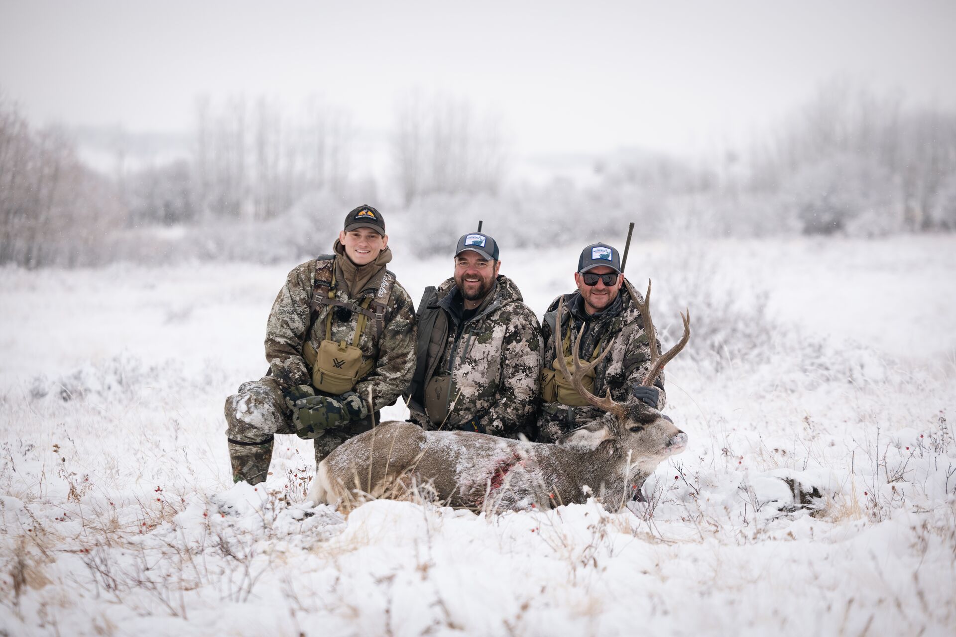 Three men sit with deer in the snow after hunt. 
