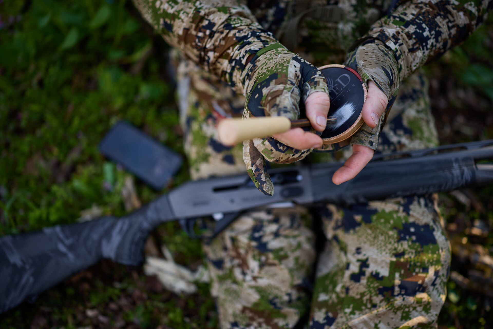 Hunter uses turkey call with shotgun nearby.