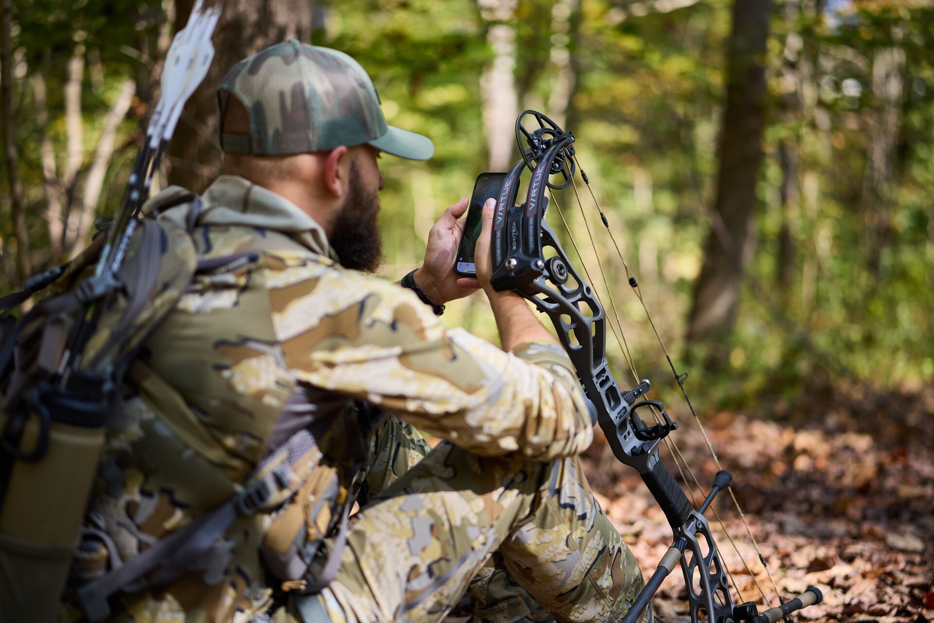 Hunter in camo on ground with bow using phone, creating a hunting plan concept.