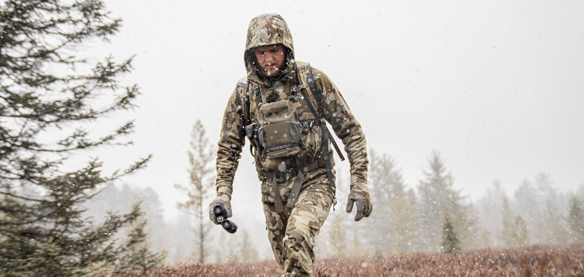 Hunter in cold weather gear walks in light snow. 